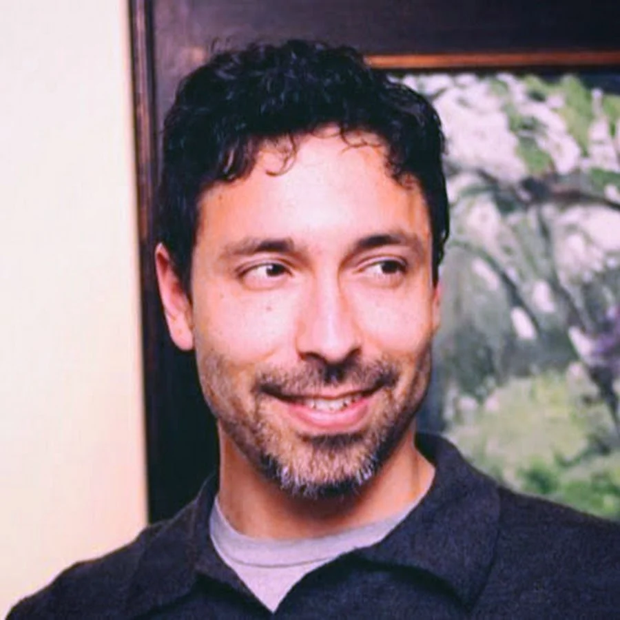 Portrait of Daniel Ibanez smiling, standing in front of one of his framed landscape paintings
